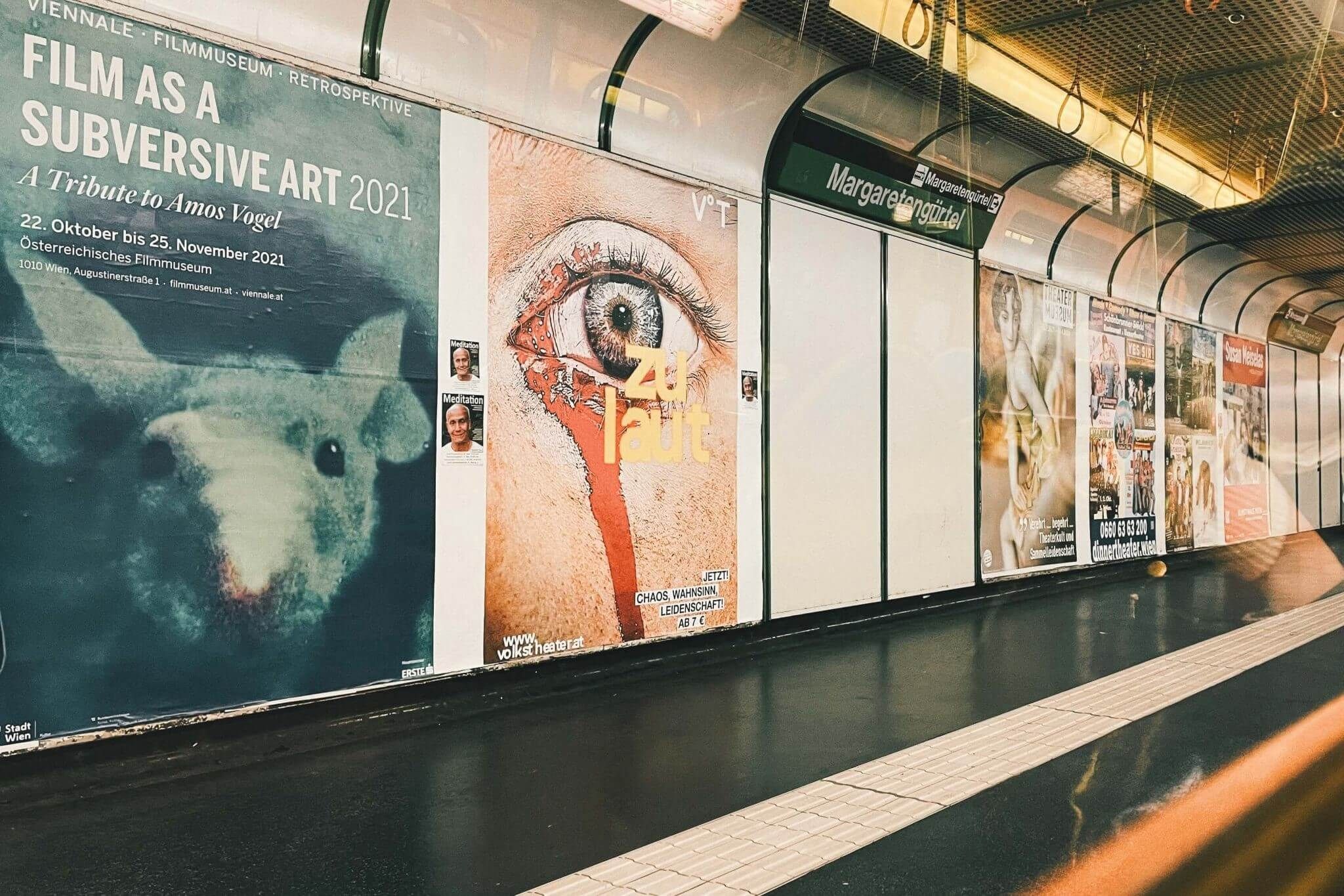 Blick auf die U-Bahn-Station Margaretengürtel in Wien mit Werbeplakaten entlang des Bahnsteigs, aufgenommen durch ein Wagonfenster.